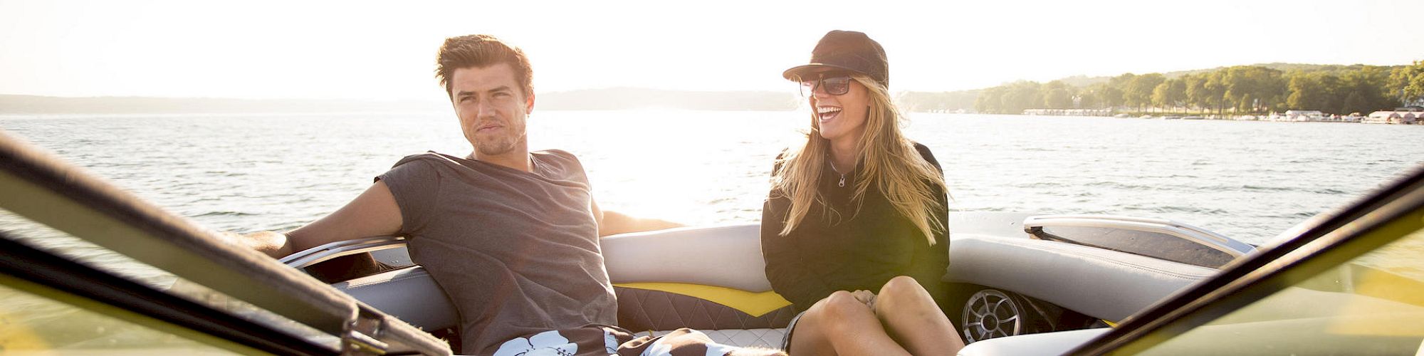 Two people enjoying a sunny day on a boat in casual attire, sitting comfortably near the front, with water and distant greenery in the background.
