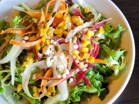 The image shows a fresh salad with mixed greens, shredded carrots, corn, onions, and small pieces of cheese in a white bowl on a dark wooden table.