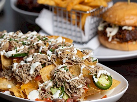 A plate of loaded chips topped with shredded meat, beans, cheese, and jalapenos, with fries, a burger, and sides in the background.