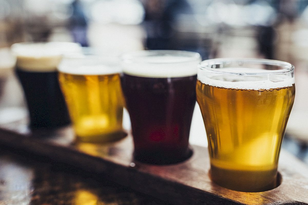 The image shows a wooden flight board with four different types of beer in glasses, each varying in color from dark to light.
