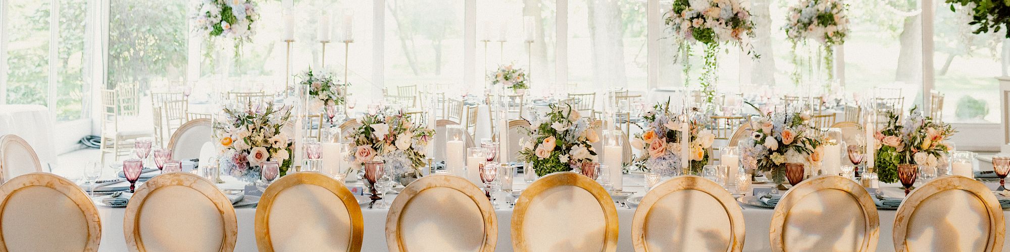 An elegant banquet setup under a white canopy with chandeliers, round-backed chairs, floral centerpieces, and table settings ready for guests.