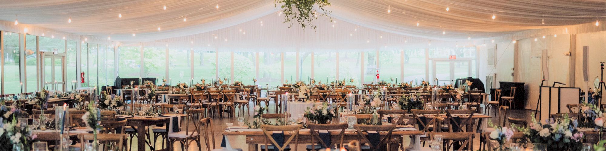 A beautifully decorated venue with draped ceilings, wooden chairs, tables set for a formal event, and hanging greenery under soft lighting.