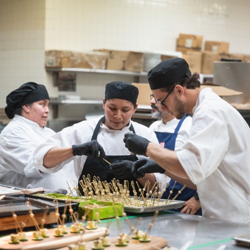 Chefs in a kitchen preparing food together, focusing on plating or garnishing dishes with a variety of small, appetizer-like servings.