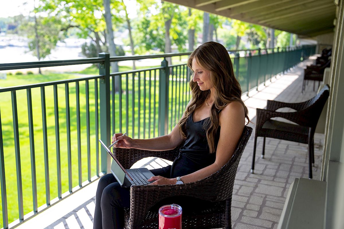 A woman sits on a balcony using a laptop, with green outdoor scenery in the background and a red tumbler on the table beside her.