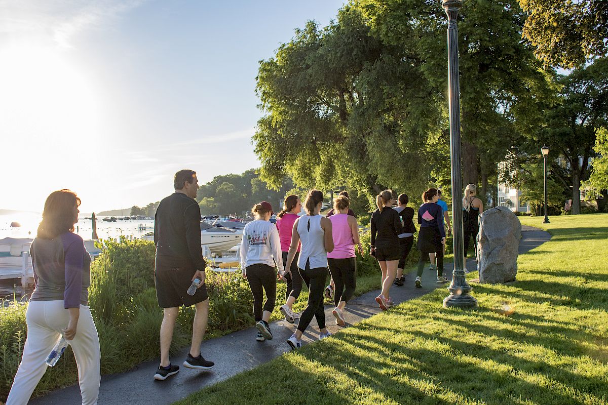 A group of people walking on a sunny pathway near the water with trees and greenery around them.