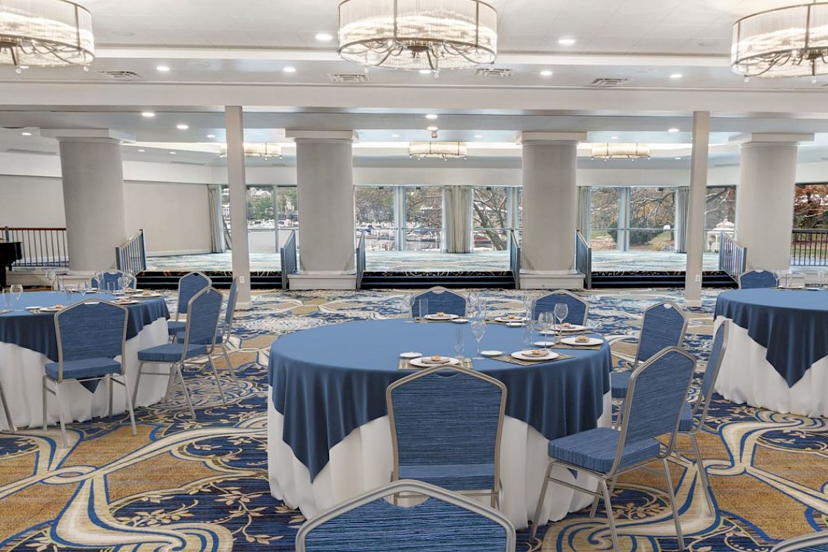 The image shows a spacious, elegantly decorated banquet hall with round tables and blue chairs, featuring chandeliers and a piano in the corner.