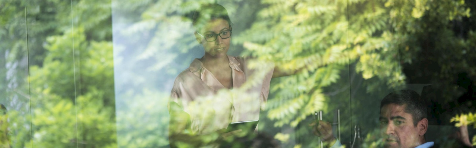 Two people are seen through a window with reflections of greenery around them, creating a layered effect in the image.