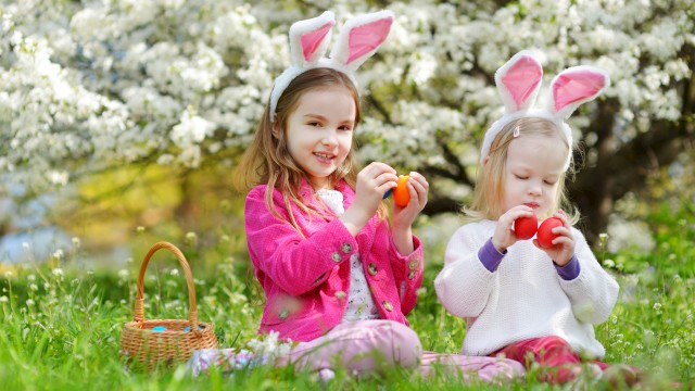 Two children with bunny ears sit on grass holding Easter eggs near a basket, with blooming trees in the background.