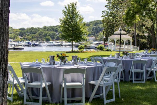 The image shows a picturesque outdoor event setup with round tables and chairs on a lawn by the water, featuring a gazebo and boats in the background.