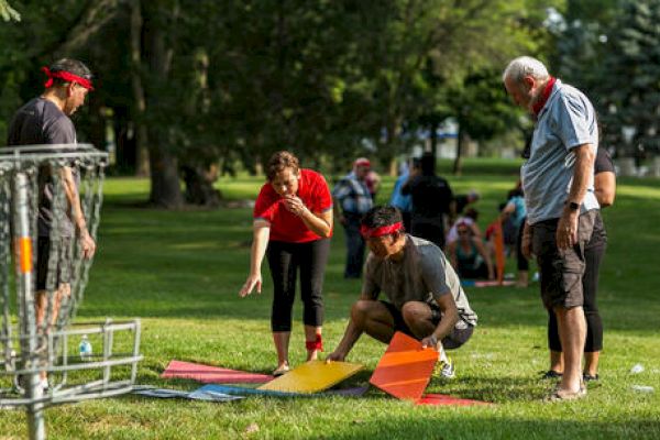 A group of people in a park, some blindfolded, appears to be engaged in an outdoor team activity involving colorful mats and a disc golf basket.