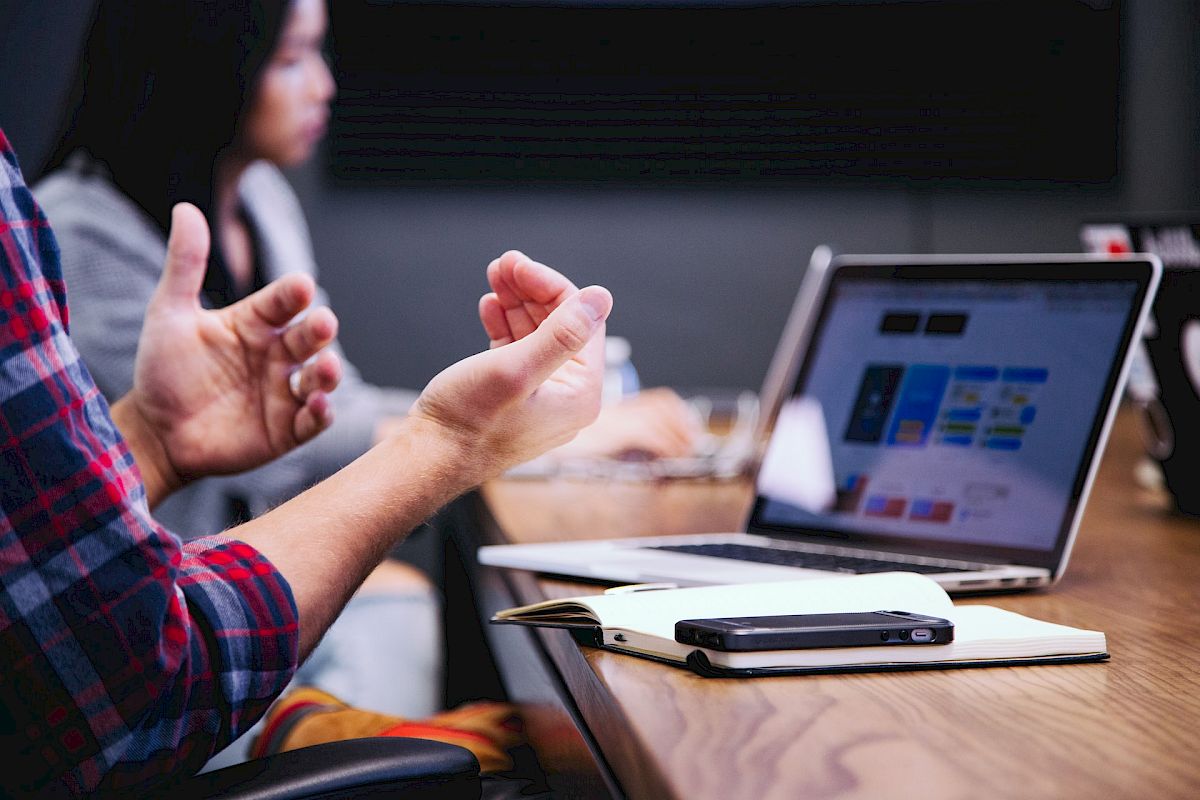 A person is gesturing during a meeting with a laptop, notebook, and smartphone on the table. Another person is blurred in the background.
