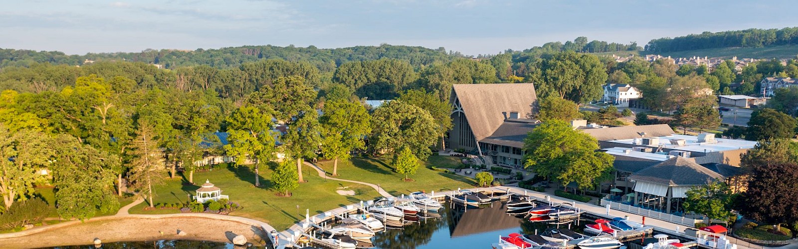 A peaceful marina with boats docked, lush green trees, and buildings in the background under a partly cloudy sky.