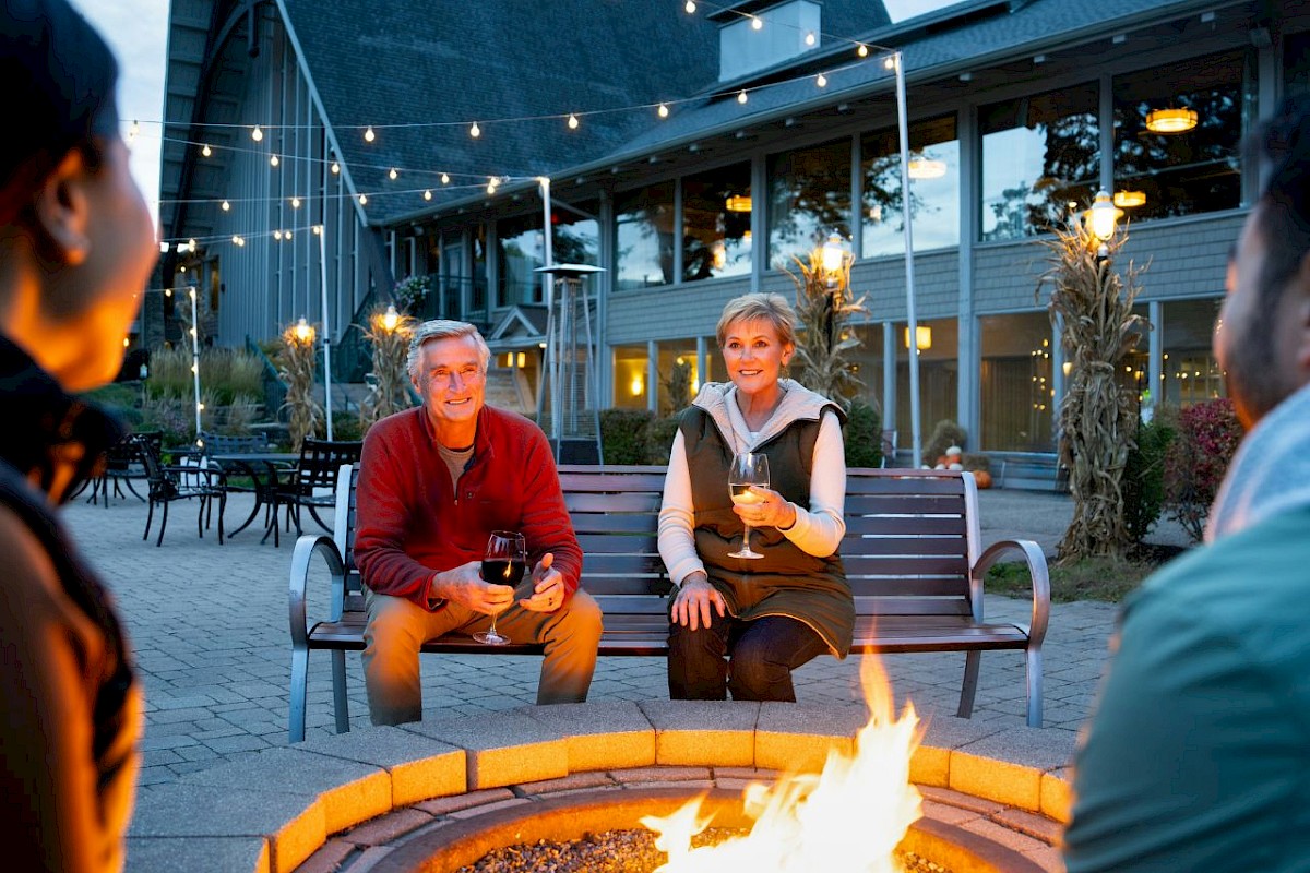 Friends enjoying evening around a fire pit with drinks, cozy atmosphere, string lights, and a modern house in the background.