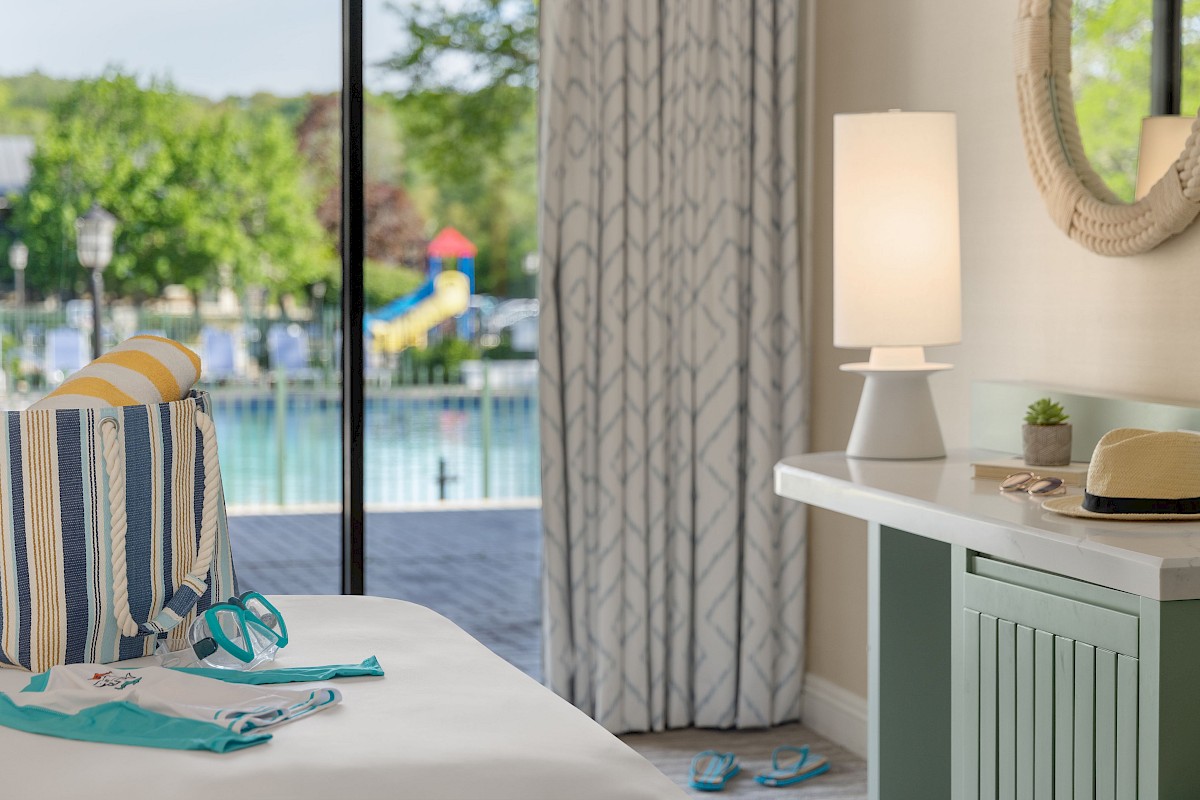 A cozy room with a view of a pool and playground outside, featuring a bed with a striped bag, swimming accessories, and summer decor.