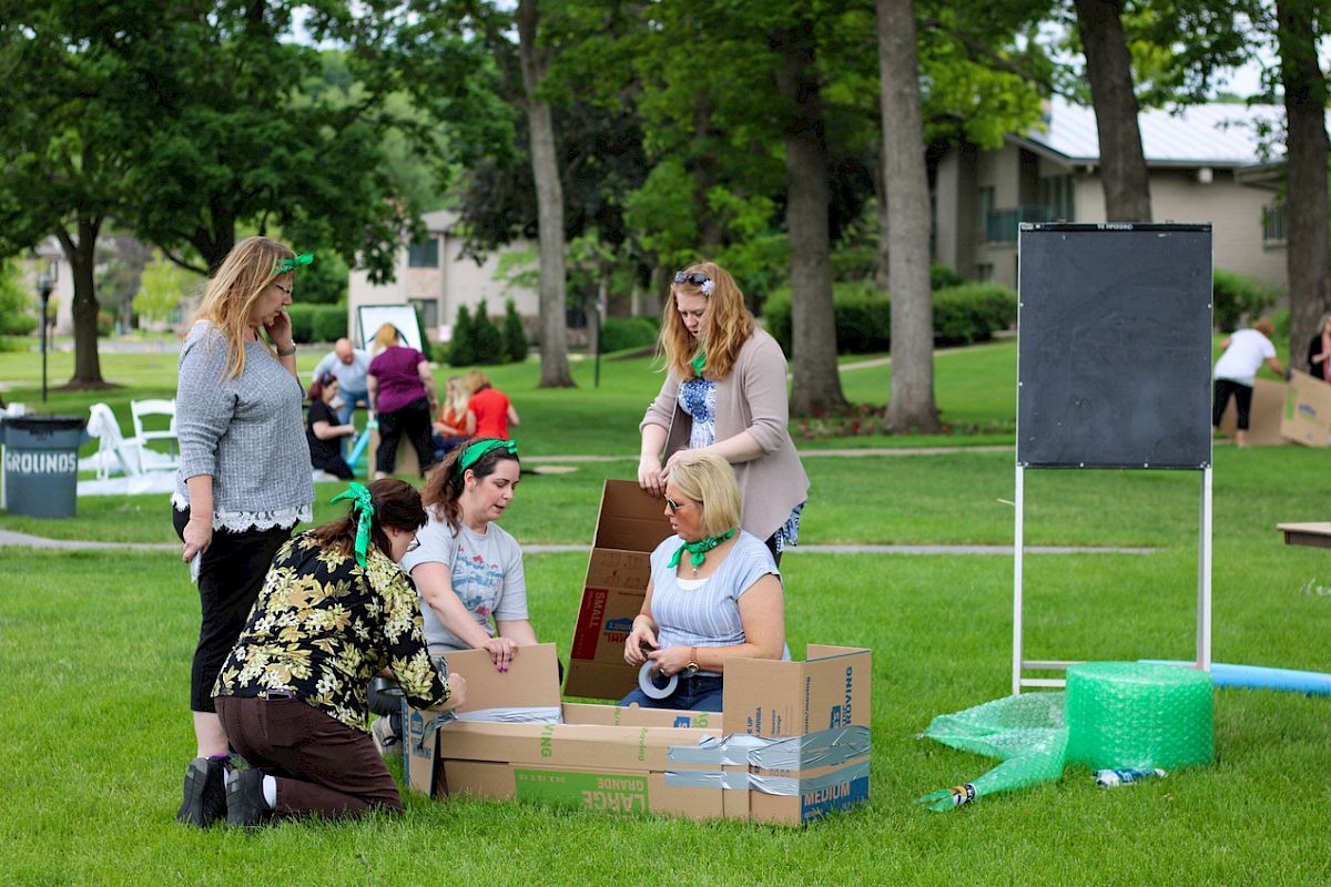 A group of people setting up cardboard boxes and bubble wrap on a grassy area, with a chalkboard in the background.