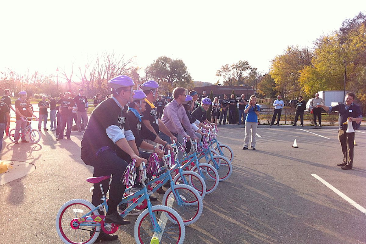 A group of adults is lined up on small bicycles wearing helmets, with an audience and trees in the background. It looks like an event.
