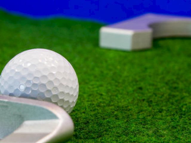 The image shows a close-up of a white golf ball on green turf next to a mini-golf putter and a corner of a mini-golf course.