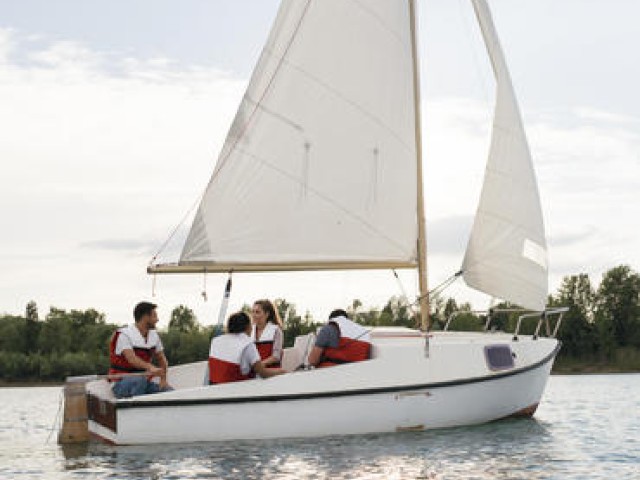 A group of four people wearing life jackets is on a white sailboat on a calm body of water with trees in the background, enjoying a leisurely ride.
