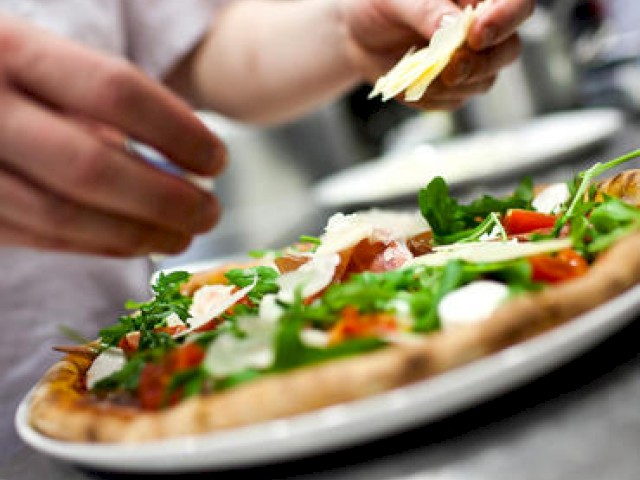 A person is placing toppings, including cheese and arugula, on a freshly made pizza, which is on a plate in what appears to be a kitchen.