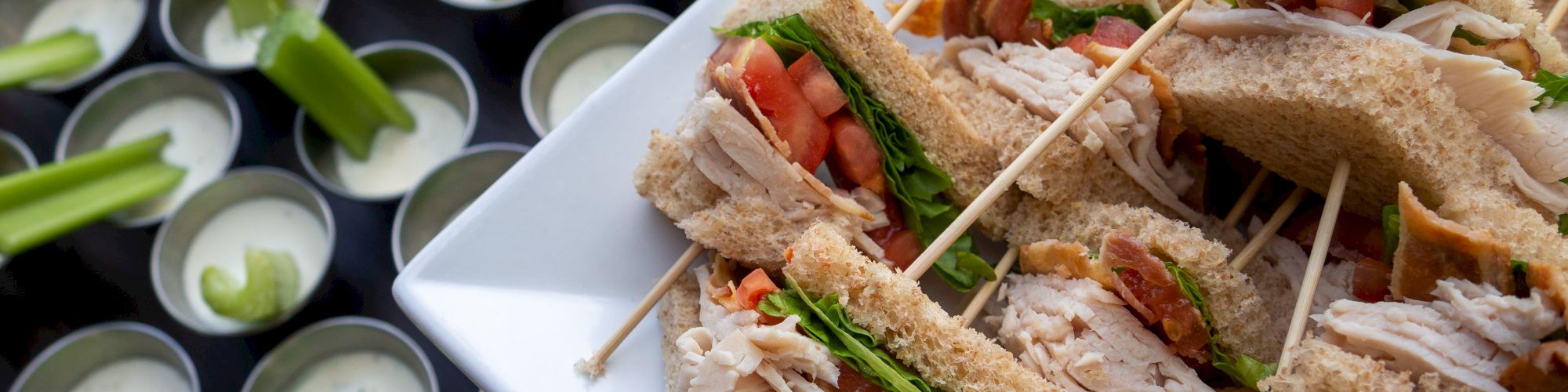 A platter of sandwiches with toothpicks, alongside cups of dipping sauce paired with celery sticks on a table, is shown in the image.