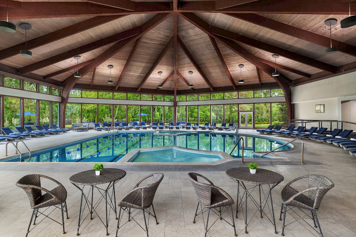 A large indoor pool area with a circular pool, wooden vaulted ceiling, and many blue lounge chairs around the water; several small tables and chairs sit near the foreground.