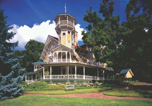 Victorian-style house with a tall tower, surrounded by greenery and a clear blue sky, featuring a charming wraparound porch.