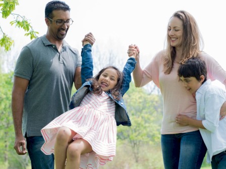 A happy family playing outside in a park, lifting a young girl on a swing and smiling.