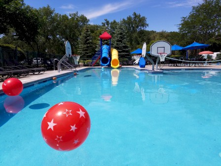 A swimming pool with floating balls, lounge chairs, waterslides, basketball hoops, and blue umbrellas on a sunny day, ending the sentence.