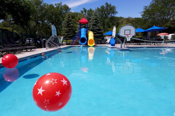 A swimming pool with floating balls, lounge chairs, waterslides, basketball hoops, and blue umbrellas on a sunny day, ending the sentence.