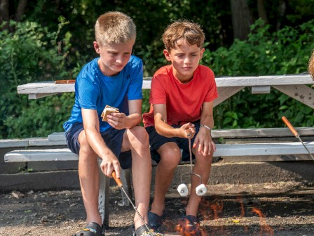 Two children are sitting by a fire pit, roasting marshmallows with sticks. They are outdoors with greenery in the background and a picnic table.