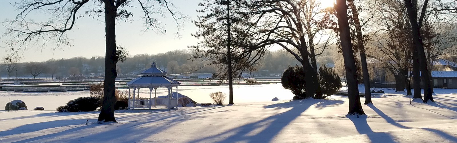 A snowy landscape with trees and a small gazebo under a clear blue sky, as the sun casts long shadows on the pristine snow.