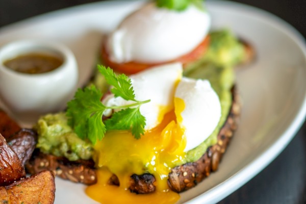 Poached eggs with avocado, cilantro, and a side of roasted potatoes on a white plate, garnished with sauce at the side.