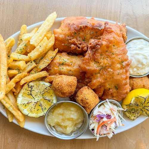 A plate of fish and chips with fries, lemon slices, sauces, coleslaw, and hush puppies on a wooden surface.