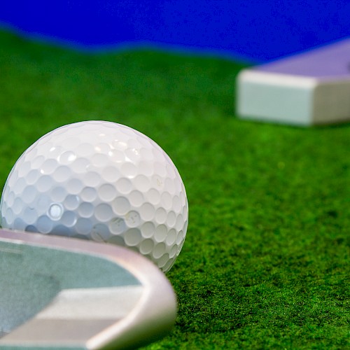A golf ball is positioned on green turf near a club, with a flagstick and hole visible in the background.