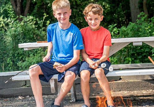 Two boys sit on a bench, roasting marshmallows over a campfire in a lush, green outdoor setting.