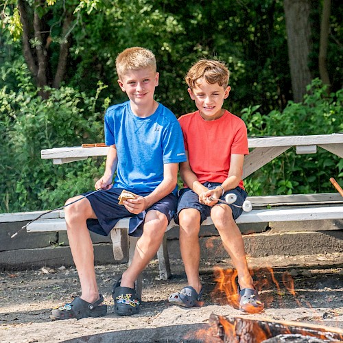 Two boys sit on a bench, roasting marshmallows over a campfire in a lush, green outdoor setting.