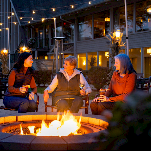Three people sit around a fire pit, enjoying drinks, with string lights and a cozy setting in the background.