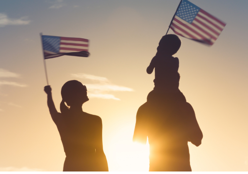 A silhouette of a person holding two American flags, with a child sitting on someone’s shoulders at sunset.