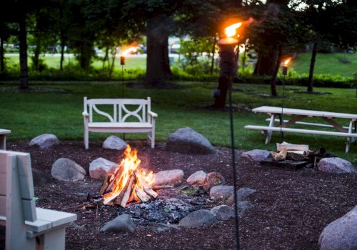 People are tending a campfire in a park-like setting with benches, torches, trees, and grassy areas.
