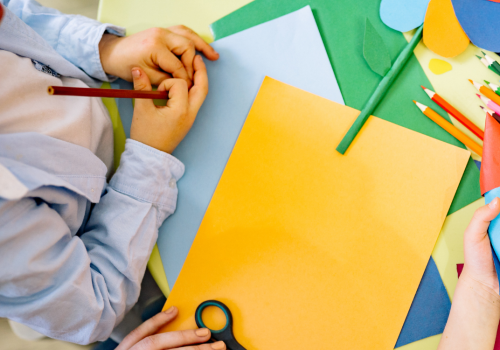 Children are making colorful paper crafts, including a rocket and a flower, using scissors, pencils, and paper on a yellow table.
