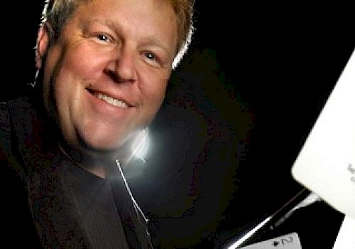 A smiling man wearing a tuxedo has playing cards and a white device in front of him, possibly demonstrating magic or a card trick.