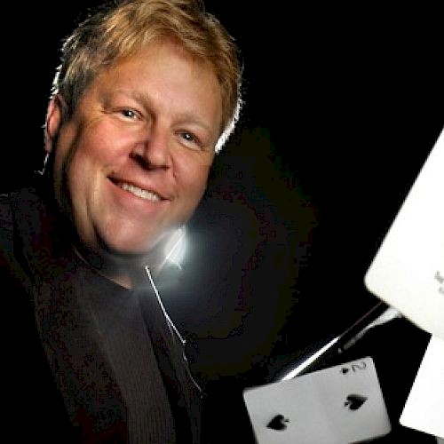 A smiling man wearing a tuxedo has playing cards and a white device in front of him, possibly demonstrating magic or a card trick.