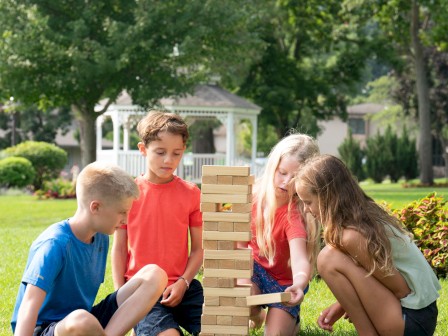 Four children are playing a giant outdoor Jenga game on a grassy lawn on a sunny day.