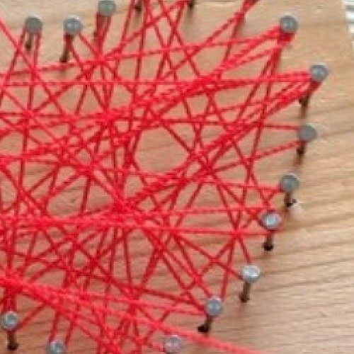 Red yarn is woven around gray nails on a wooden surface, creating a complex, interconnected string art pattern.