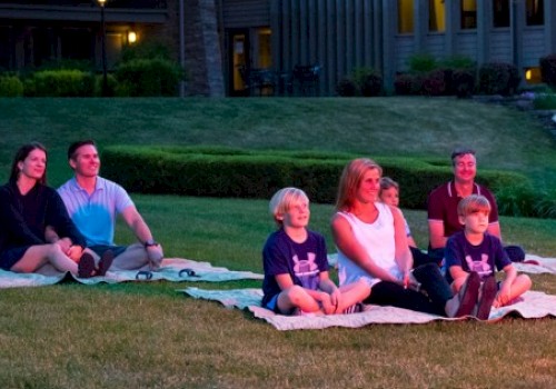 A family is sitting on blankets outdoors, watching an evening event on the grass in front of a residential area.
