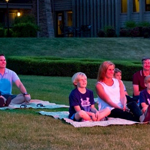 A family is sitting on blankets outdoors, watching an evening event on the grass in front of a residential area.