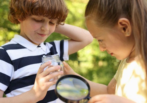 A boy and girl are exploring something in a jar outdoors, using a magnifying glass, amid lush greenery.