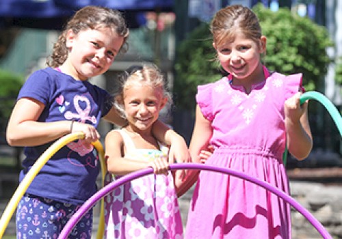 Three smiling young girls hold hula hoops and stand outdoors, enjoying a sunny day at a park or playground.