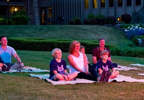 A family is sitting on blankets outdoors, watching an evening event on the grass in front of a residential area.