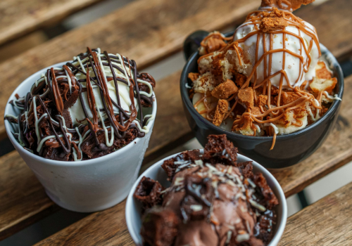 Three bowls of decadent ice cream with various toppings are displayed on a rustic wooden table.